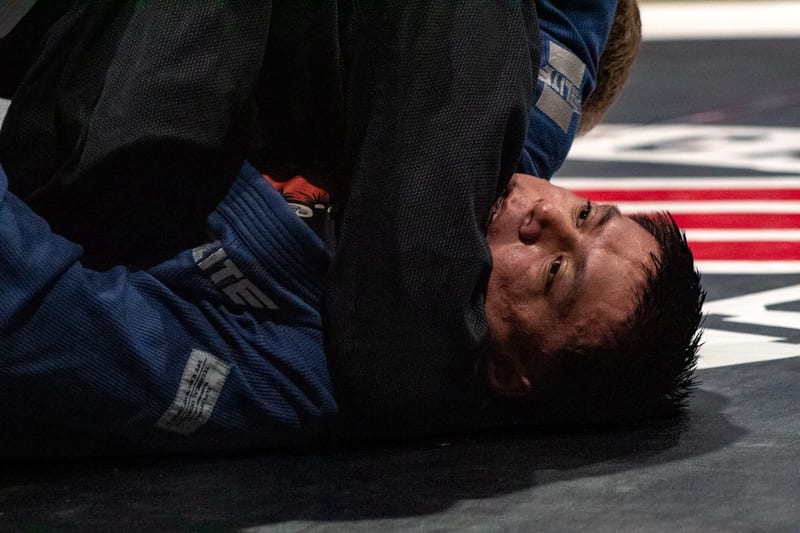 A competitor in a blue gi defends from the bottom with the match clock running overhead.