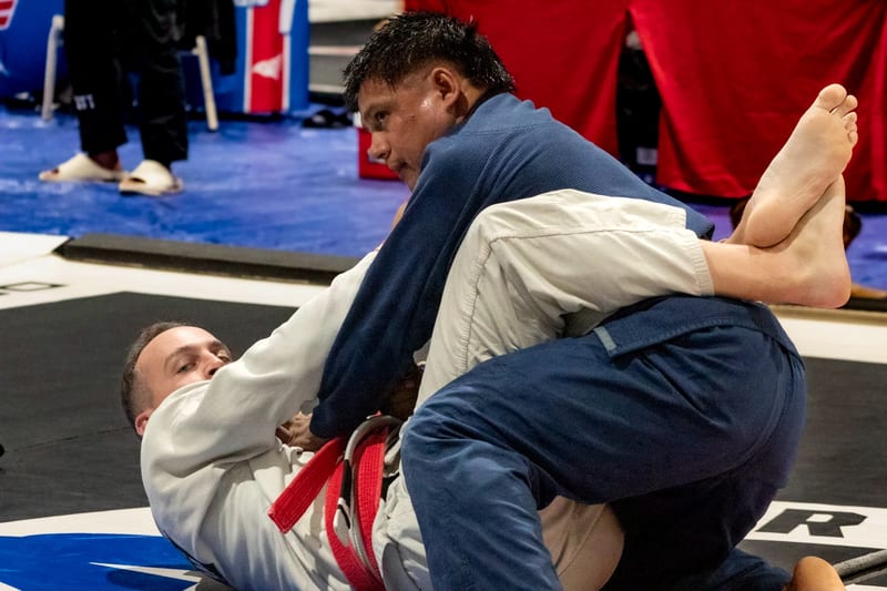 A competitor in a blue gi works from top position over his opponent during a gi match.