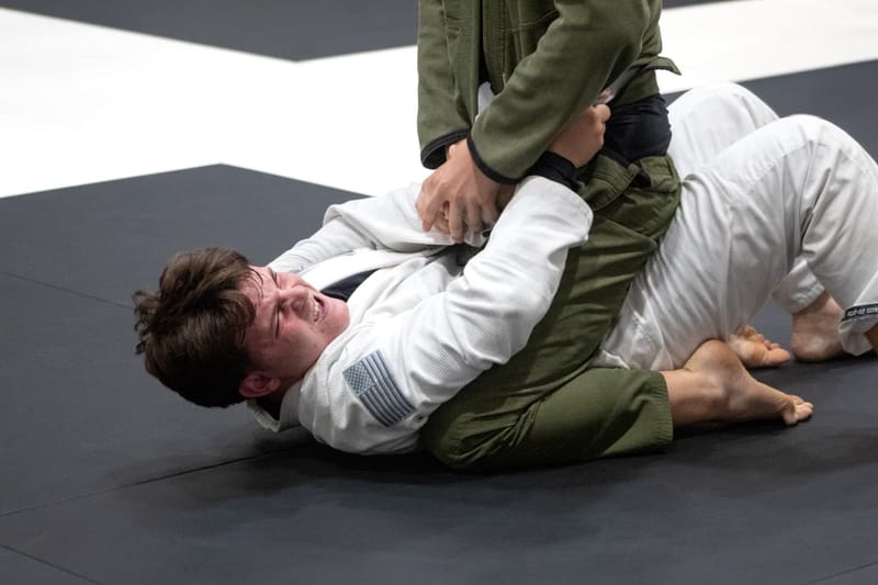 An adult in a white gi grimaces while defending on the bottom during a grappling match.