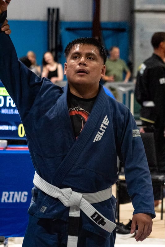 A competitor in a blue gi has his hand raised in victory in front of the scoreboard.