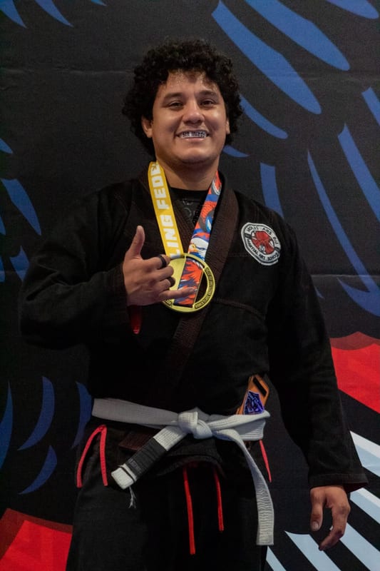 A smiling adult competitor in a black gi shows off his gold medal in front of the tournament banner.