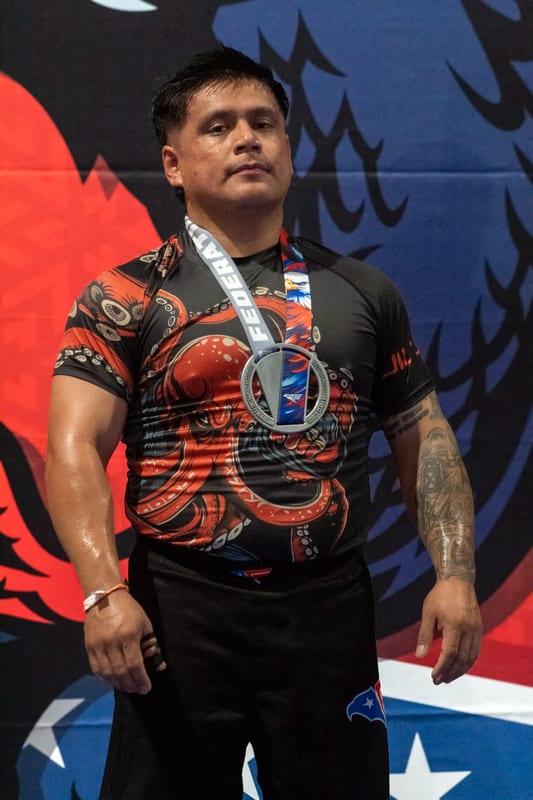 An adult competitor in an octopus rashguard wears his medal in front of the tournament banner.