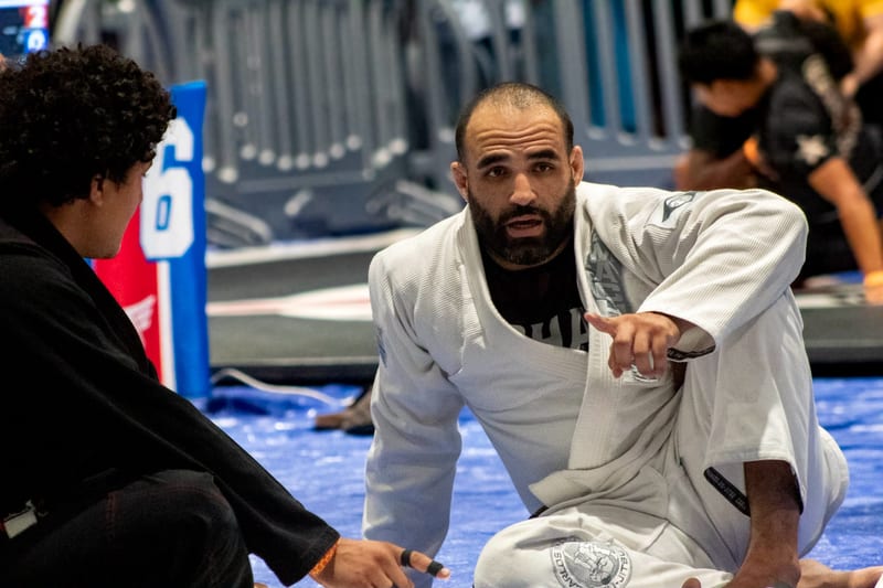 A competitor in a white gi kneels at the mat edge between matches.