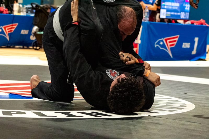 An adult competitor in a black gi works from top position during a gi match.