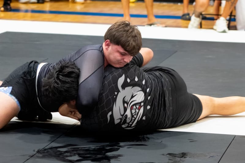 An adult grappler in a grey rashguard applies side-control pressure during a no-gi match.