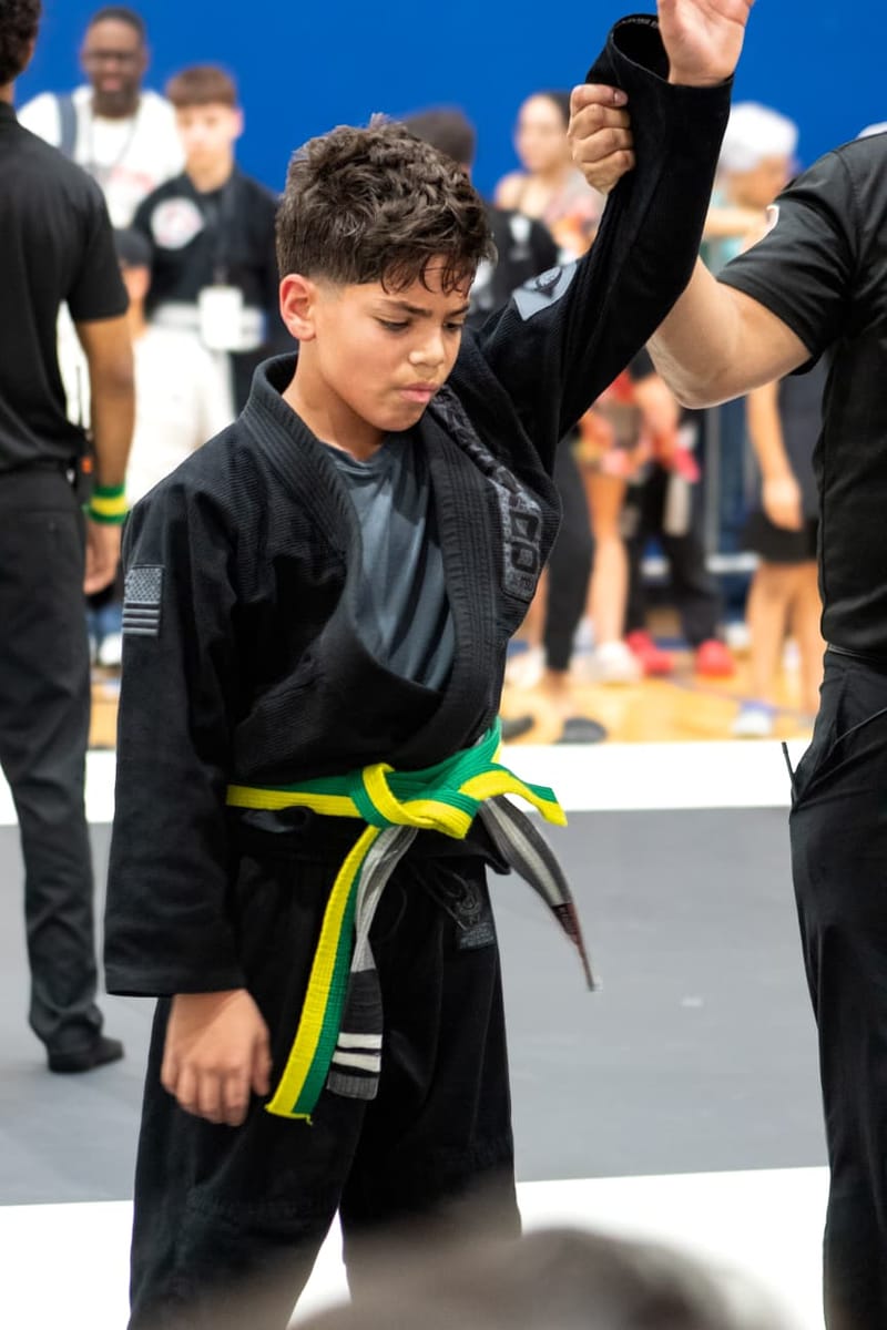 A boy in a black gi with a green-and-yellow belt has his arm raised by a referee after a win.