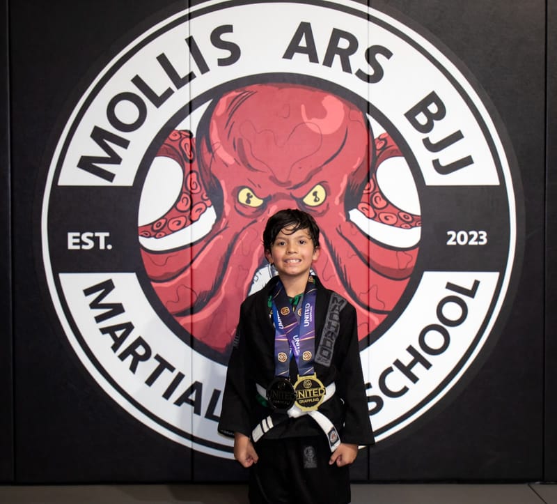A smiling boy in a black gi stands wearing silver and gold medals at the Mollis Ars BJJ logo wall.