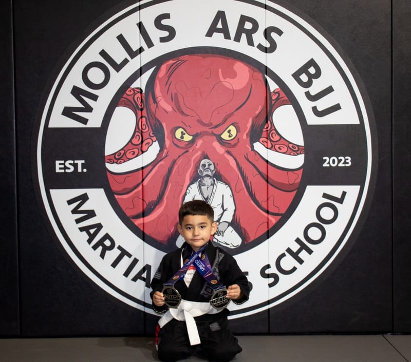 A dark-haired boy in a black gi kneels holding two silver medals in front of the octopus logo wall.