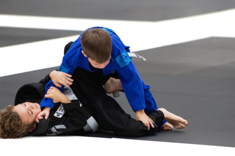 A boy in a blue gi passes the guard and takes side control over an opponent in a black gi.
