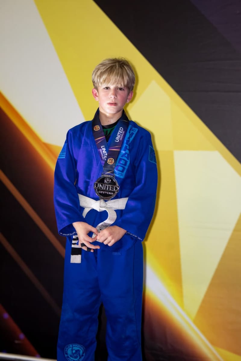A boy in a blue gi poses with a silver medal around his neck against a bright graphic backdrop.