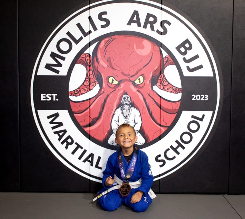 A smiling boy in a blue gi kneels wearing a bronze medal at the Mollis Ars BJJ logo wall.