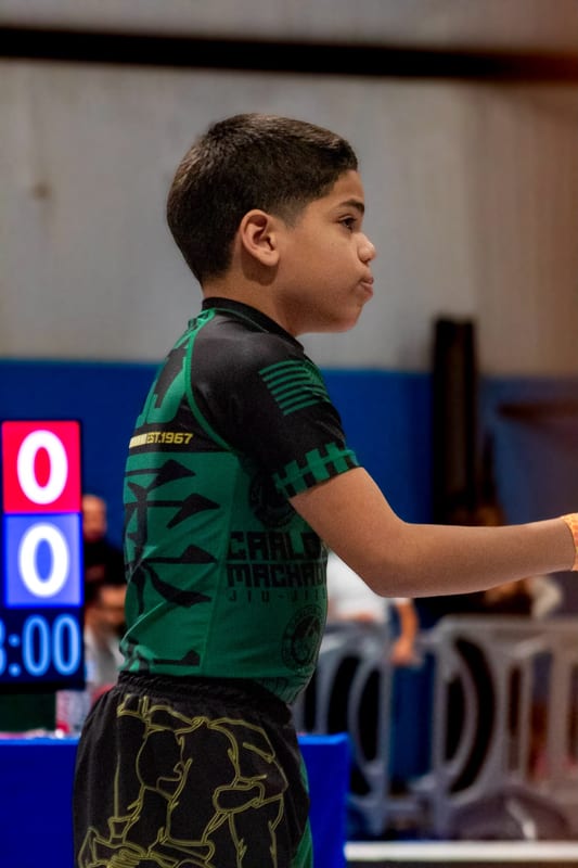 A boy in a green and black rashguard walks onto the mat beside the scoreboard.