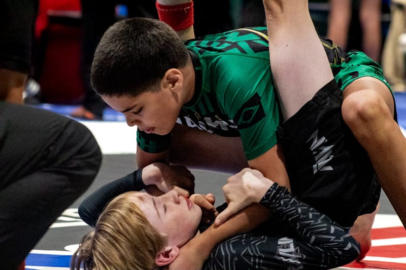 A boy in green and black holds top position over his opponent during a kids no-gi match.