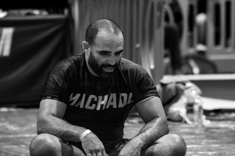 Black-and-white photo of a coach in a Machado rashguard smiling from the side of the mat.