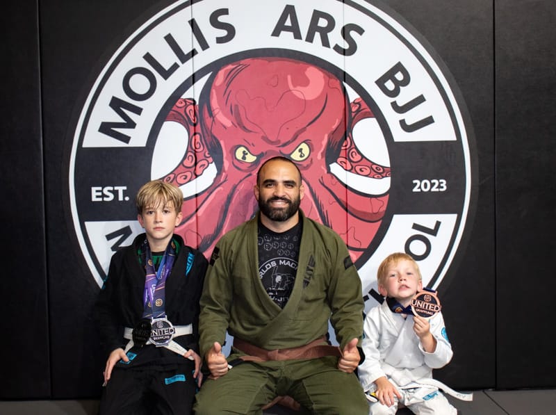 A brown-belt coach sits between two young students in gis holding their medals at the logo wall.