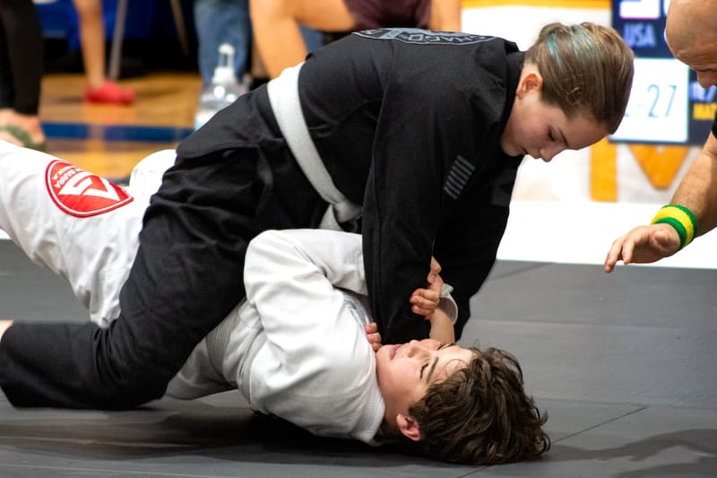 A girl in a black gi holds the mount over an opponent in a white gi during a tournament match.