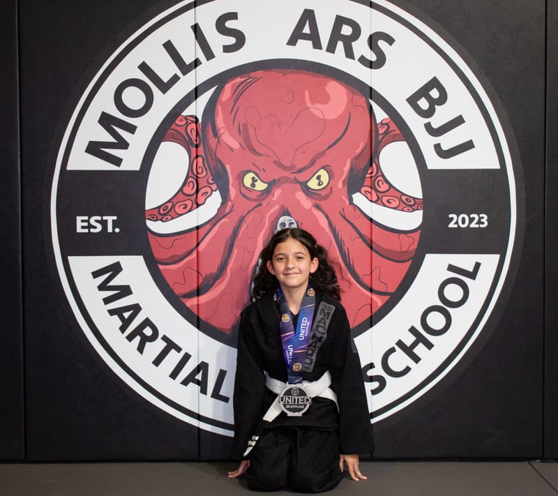 A girl with pigtails in a black gi kneels wearing a silver medal at the Mollis Ars BJJ logo wall.