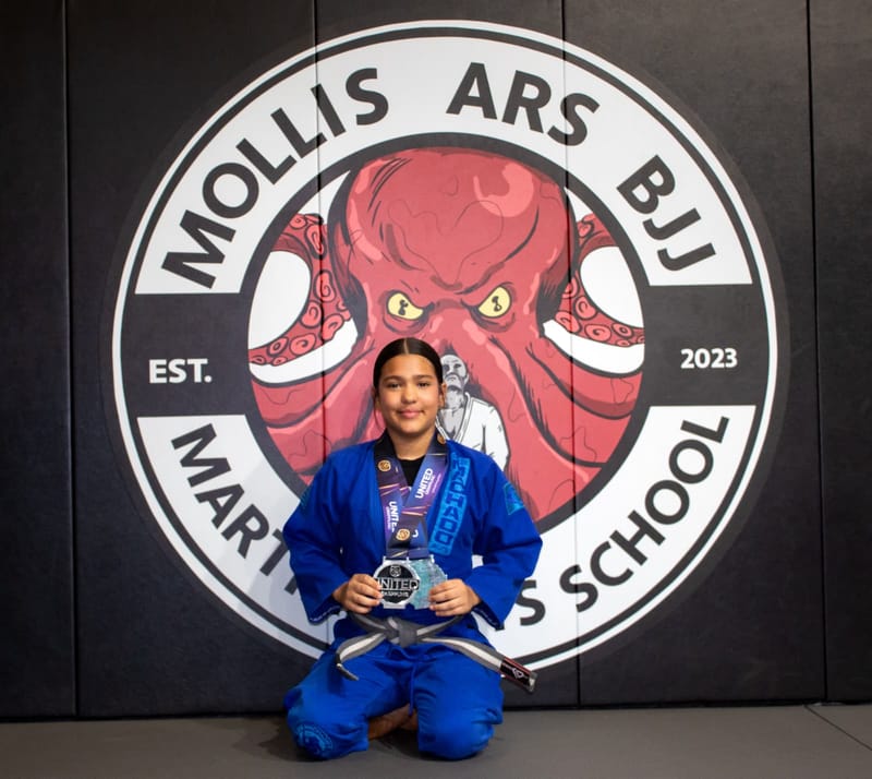 A girl in a blue gi kneels holding a clear United Grappling award in front of the Mollis Ars BJJ octopus logo.