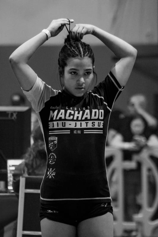 Black-and-white photo of a focused girl in a Machado rashguard with her hands on her head before a match.