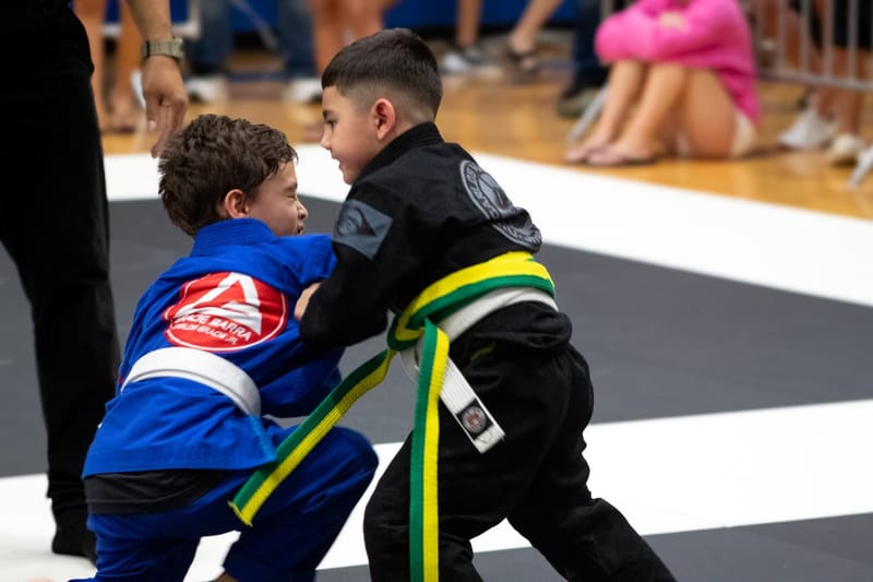 A girl in a purple gi controls her opponent on the mat as a referee watches during a match.