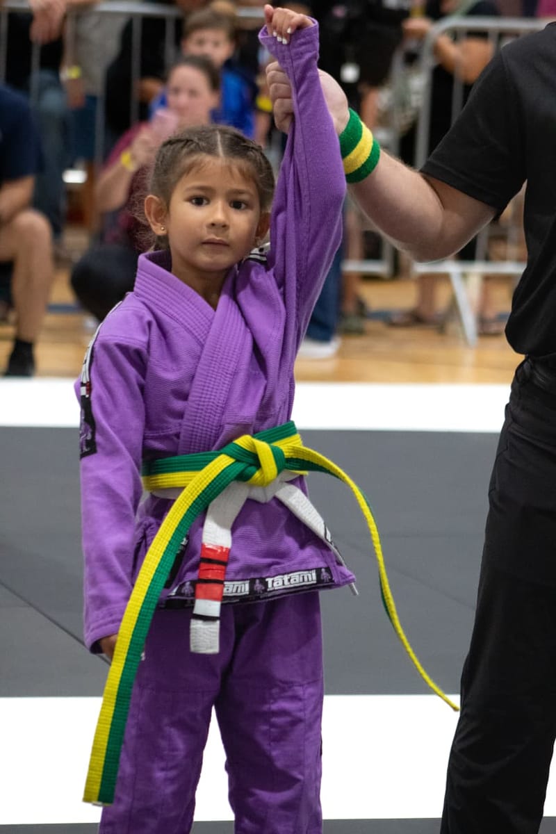 A young girl in a purple gi with a green-and-yellow belt has her arm raised by a referee after winning her match.