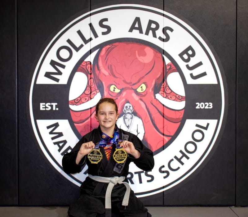 A grinning girl in a black gi shows off two gold medals in front of the Mollis Ars BJJ logo wall.