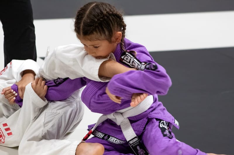 A young girl in a purple gi controls her opponent in a white gi during a jiu-jitsu match.