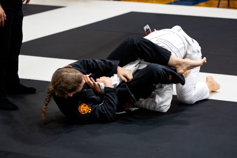 A girl in a black gi works an attack from the bottom during a competition ground exchange.