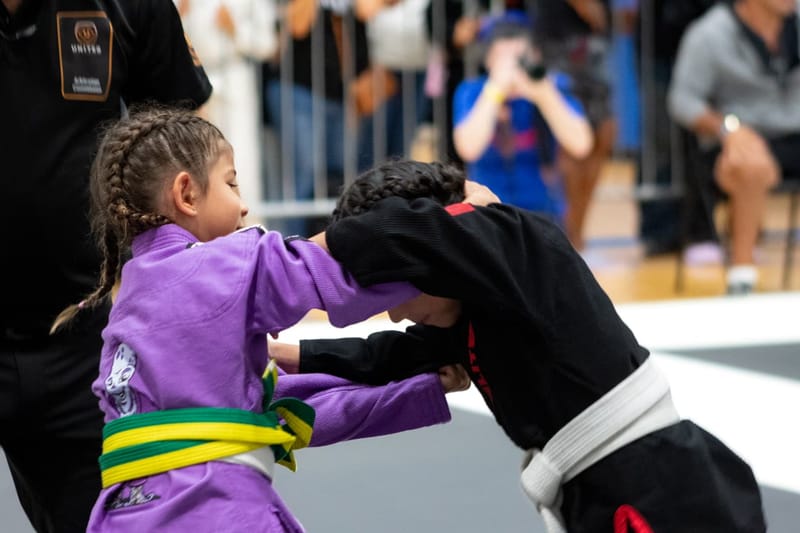 A girl in a purple gi and a girl in a black gi fight for grips in a standing clinch during a match.
