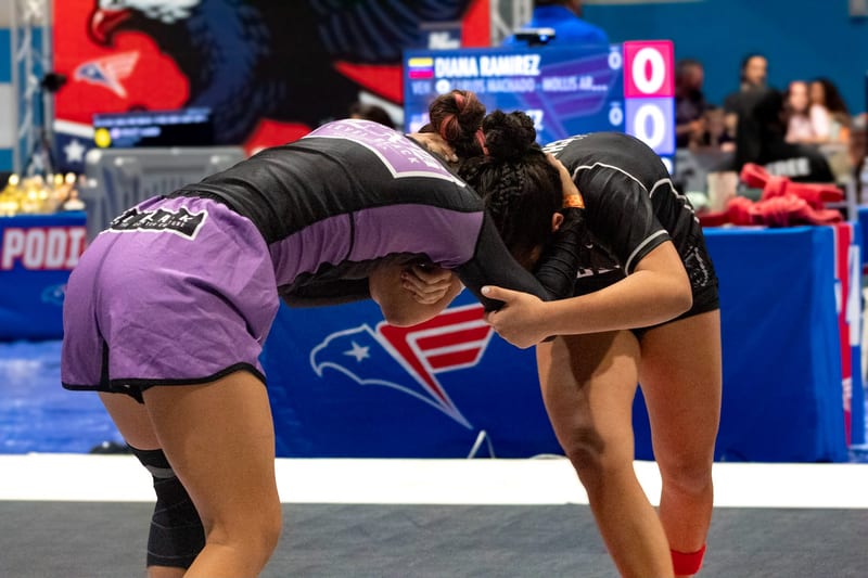 Two girls lock up at the start of a no-gi match in front of the scoreboard.