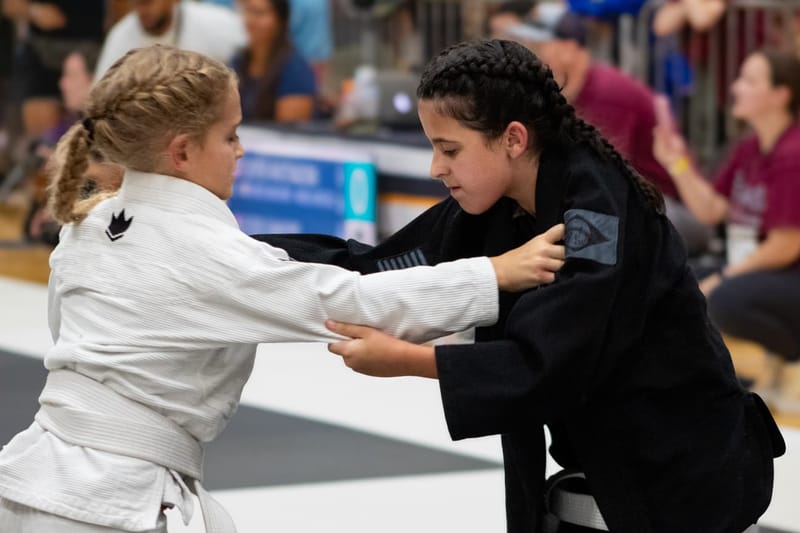 Two girls in gis battle for grips while standing during a jiu-jitsu competition match.