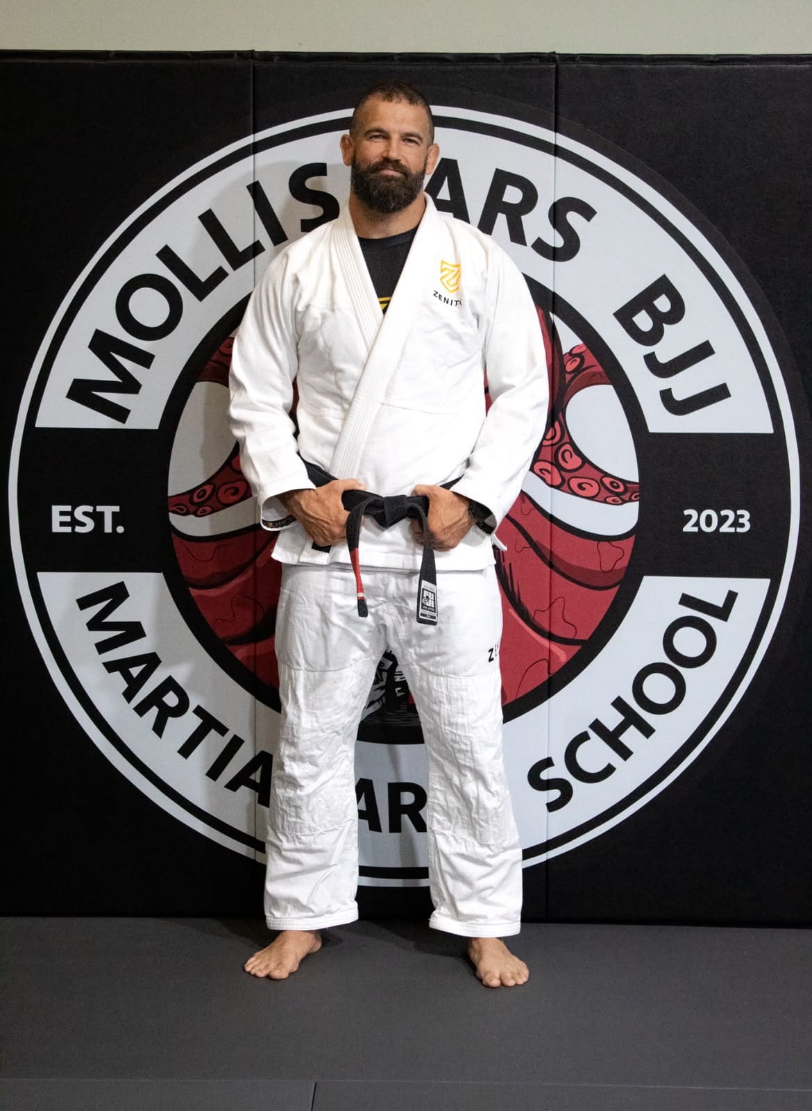 Jeremy Reid, black belt instructor at Mollis Ars BJJ, in his gi holding his black belt in front of the academy logo wall