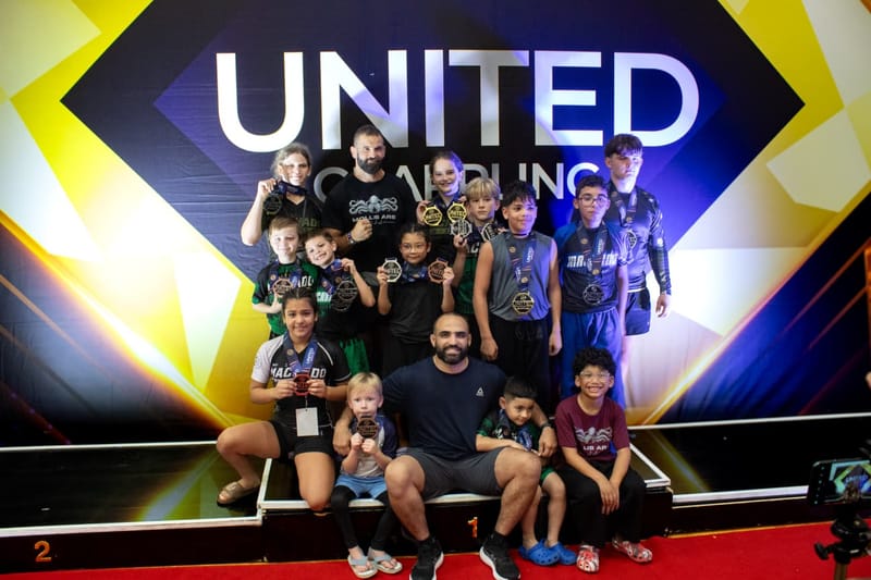 A group of young athletes and their coaches hold up medals together in front of a large tournament banner.