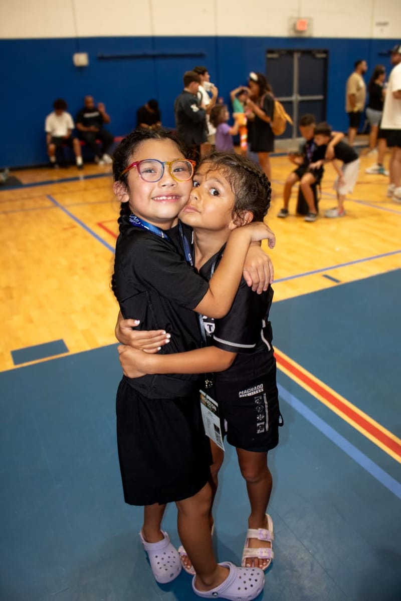 Two young teammates share a hug in the gym, one planting a kiss on the other’s cheek.