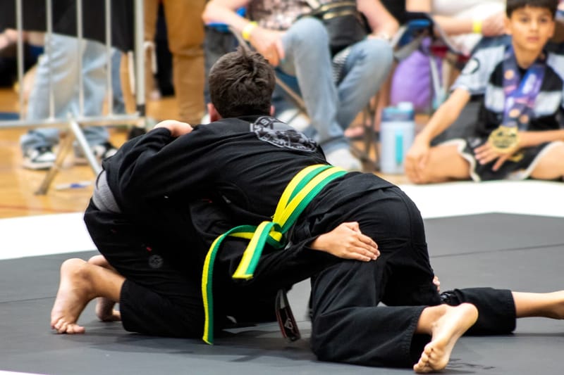 Two young boys in black gis clinch on the mat as a medaled child watches from the sideline.
