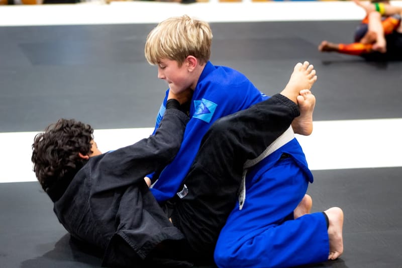 A blond boy in a blue gi controls an opponent in a black gi from the top during a kids match.