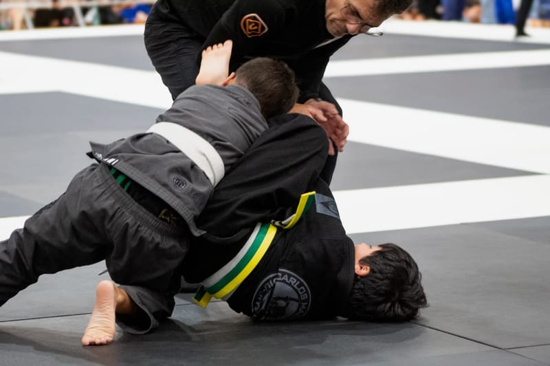 Two boys grapple on the mat while a referee leans in to watch during a tournament match.