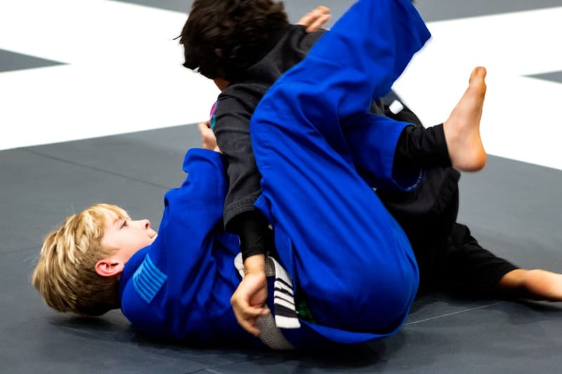 A boy in a blue gi controls an opponent from guard during a kids jiu-jitsu match.