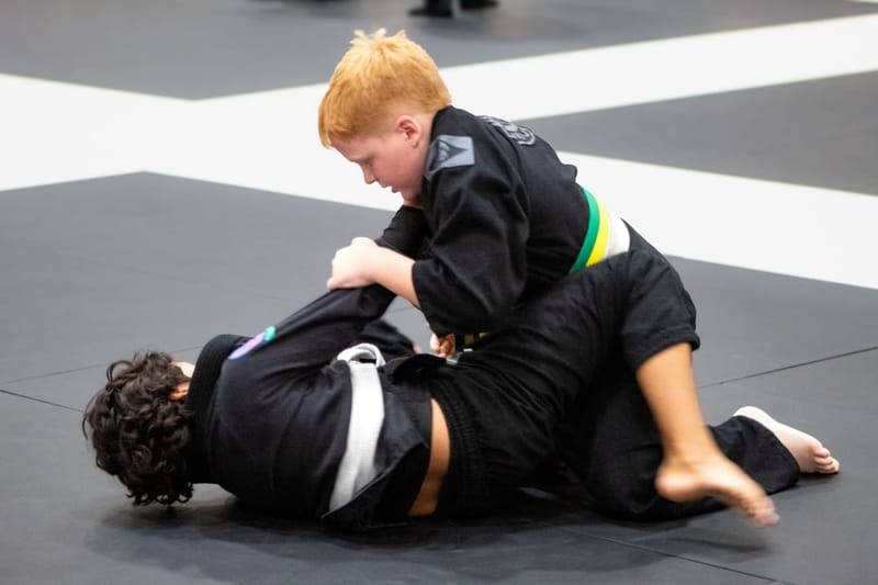 A red-haired boy in a black gi works to pass his opponent’s guard during a ground exchange.