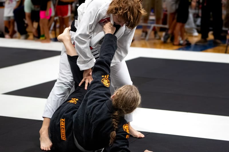 A child in a white gi stands to pass while an opponent plays open guard from the mat during a match.