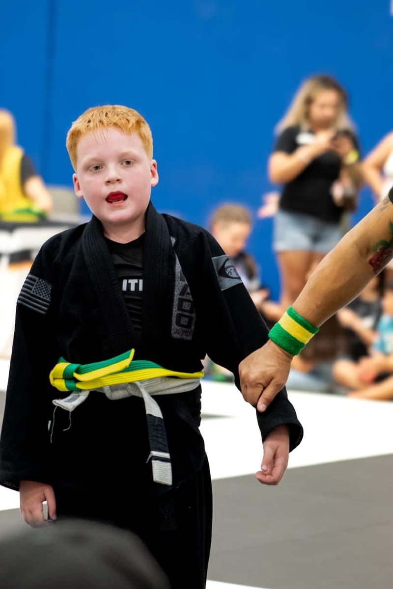 A red-haired boy in a black gi stands beside a referee holding his arm after a match.