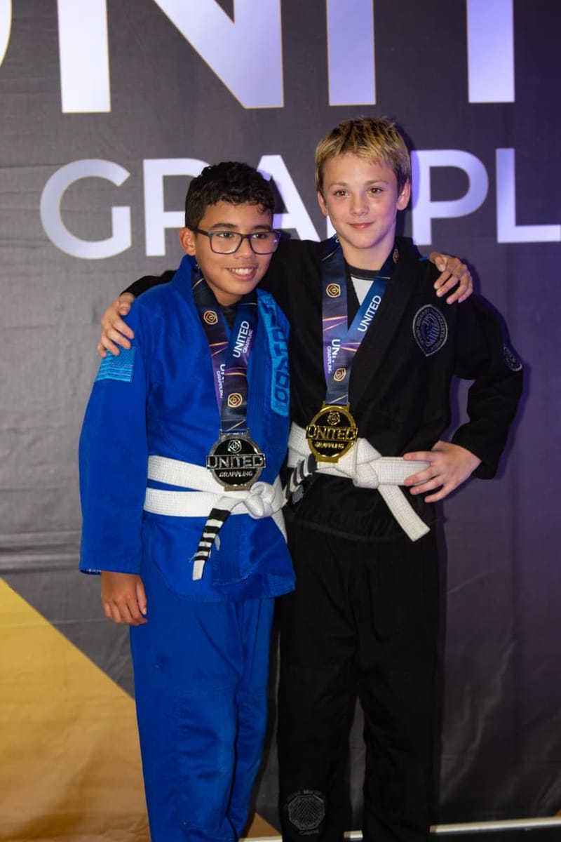 Two boys in blue and black gis pose with gold medals in front of a tournament backdrop.