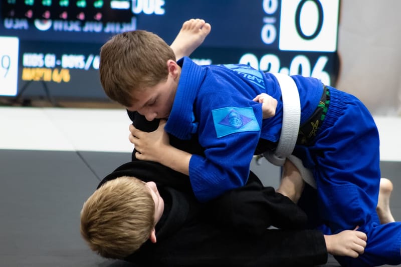 A boy in a blue gi controls his opponent on the mat in front of a match scoreboard.