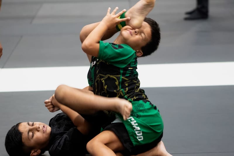 Two young boys scramble during a no-gi jiu-jitsu match, one in a green rashguard defending on top.