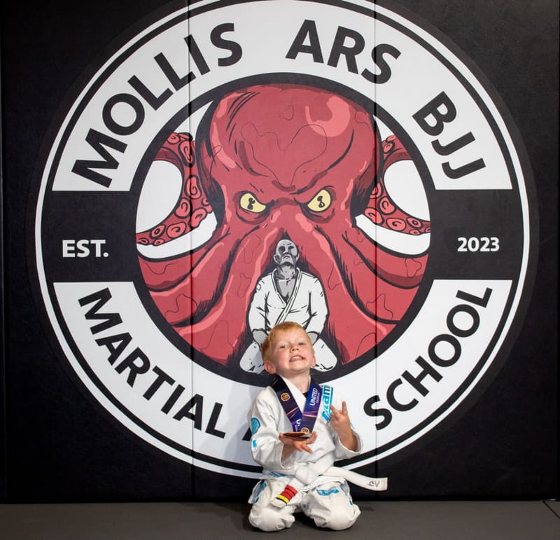 A grinning little boy in a white gi kneels with a medal in front of the Mollis Ars BJJ logo wall.