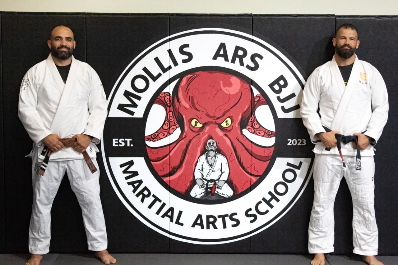 Dhavid Antunez and Jeremy Reid, the coaching staff at Mollis Ars BJJ, standing in front of the academy logo wall