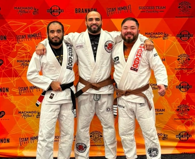 Three Mollis Ars BJJ athletes in white gis, one black belt and two brown belts, standing arm in arm in front of the Carlos Machado Jiu-Jitsu banner