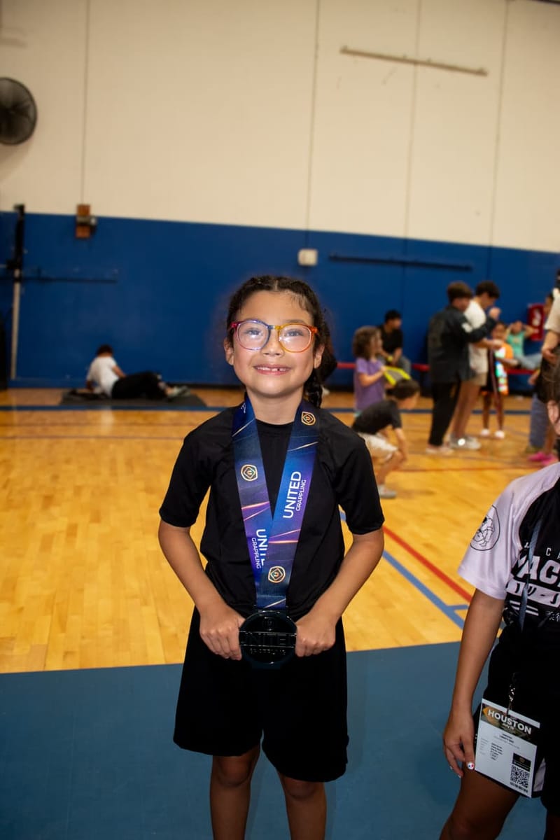 A smiling young girl wearing glasses holds up her United Grappling medal in the gym.