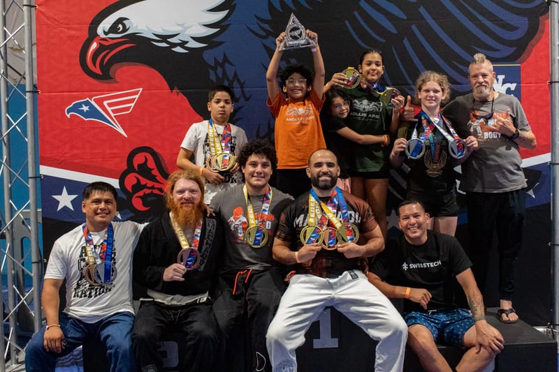 The Mollis Ars BJJ team poses together with medals and a trophy in front of the tournament banner.
