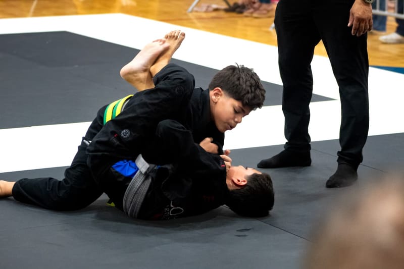 Two teenage competitors in black gis battle for position on the mat during a jiu-jitsu match.